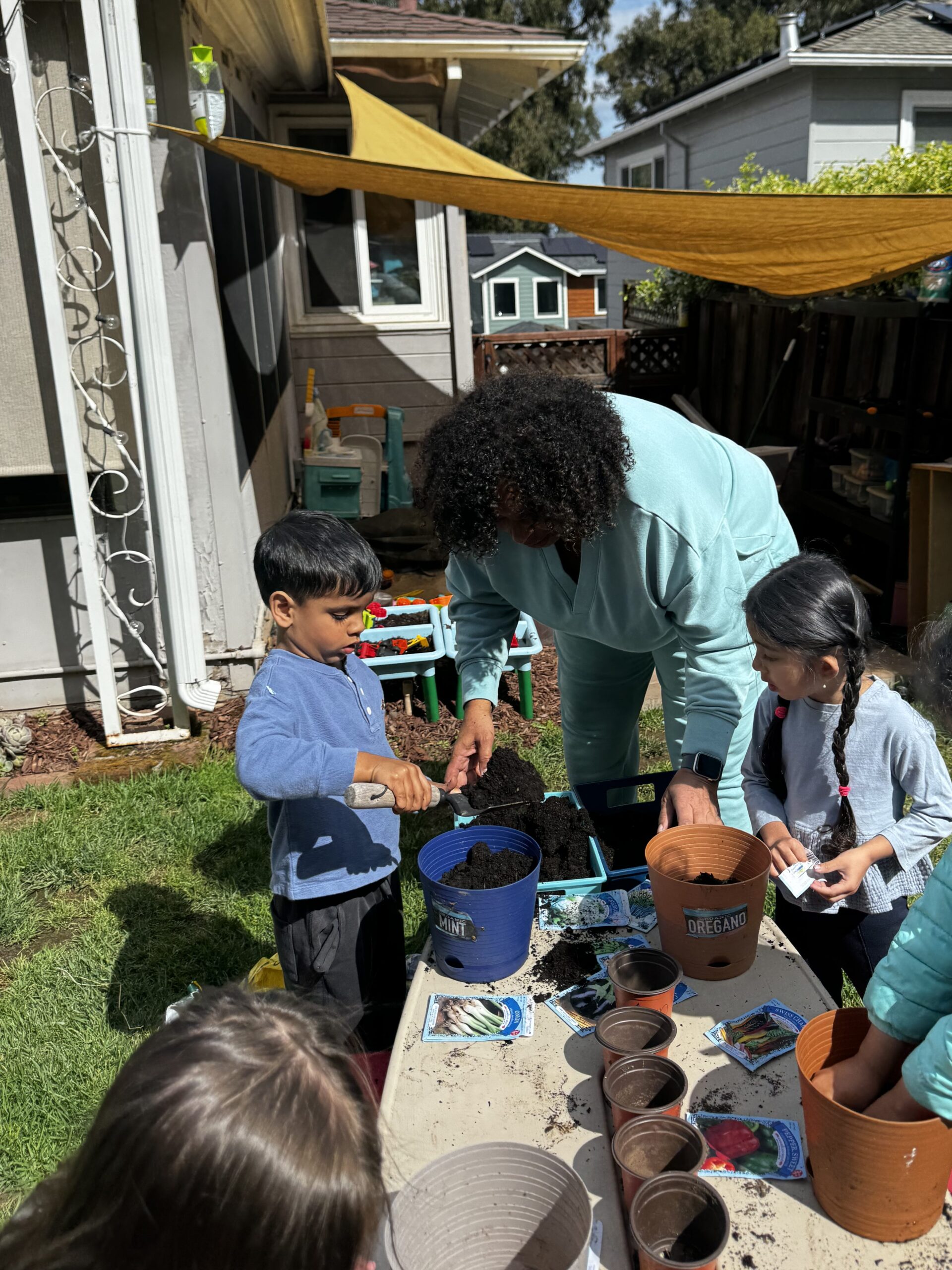 An adult and children fill plant pots with soil at a table outside on a sunny day.