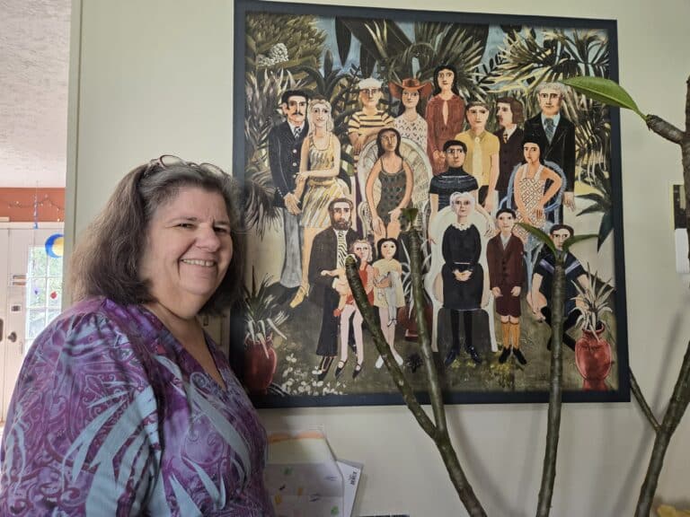 Smiling woman in a purple shirt stands by a colorful painting of many people in a garden setting.