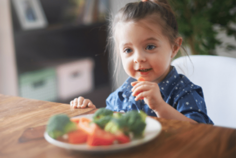Young girl sitting at a table, smiling and holding a carrot stick, with a plate of vegetables in front of her.