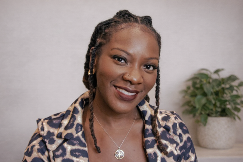 Woman with braided hair smiles, wearing a leopard-print shirt and necklace; plant in background.