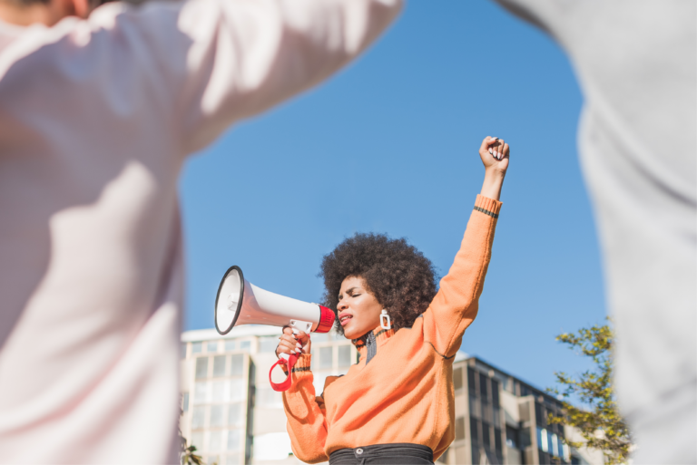 A woman with a megaphone raises her fist while protesting outdoors under a clear blue sky.