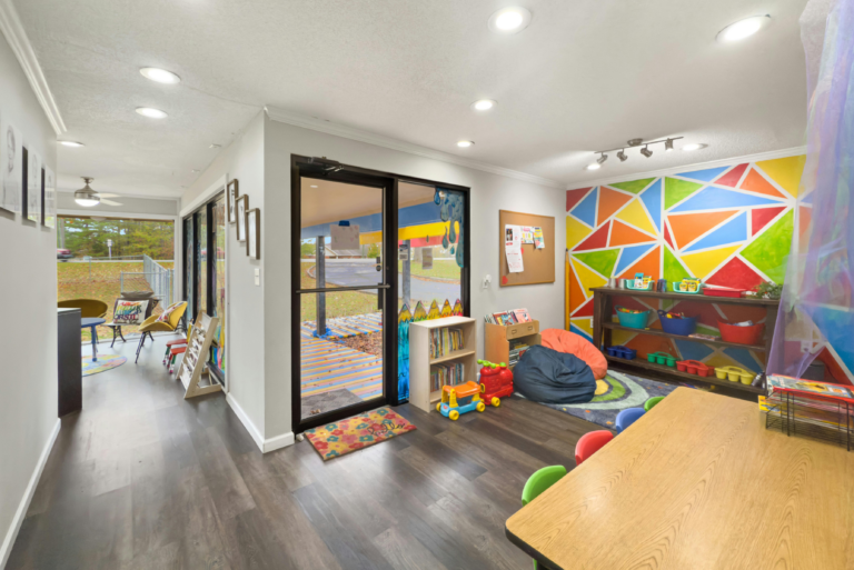 Colorful children's playroom with geometric mural, toys, bookshelves, and a table with small chairs.