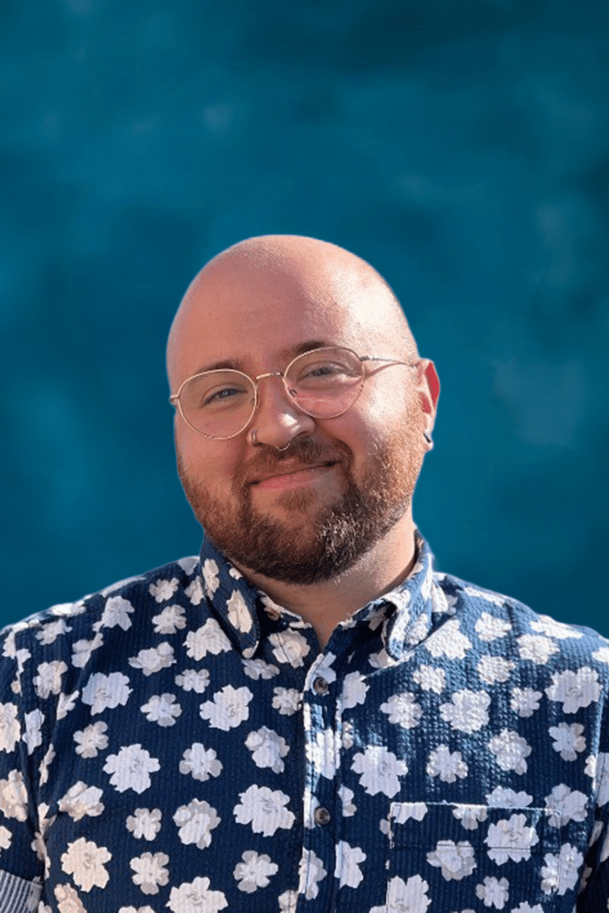 Smiling person with glasses and a floral shirt stands in front of a bright blue background.