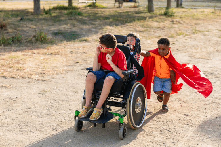 Three kids play outdoors, one in a wheelchair, while another pushes and both wear red capes.