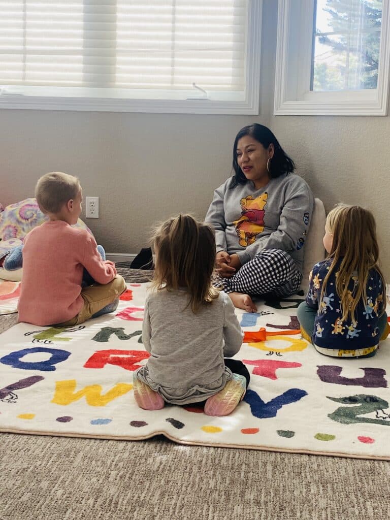 A teacher sits on an alphabet mat with three young children, engaging in a group activity in a bright room.