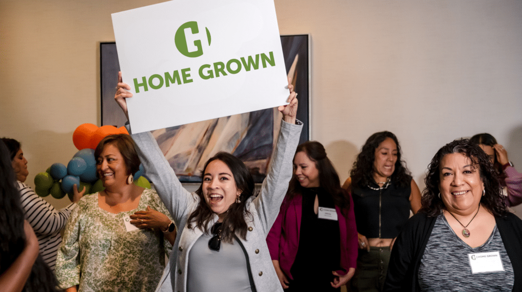 Smiling woman holds "Home Grown" sign above her head; a group of women stand and smile behind her.