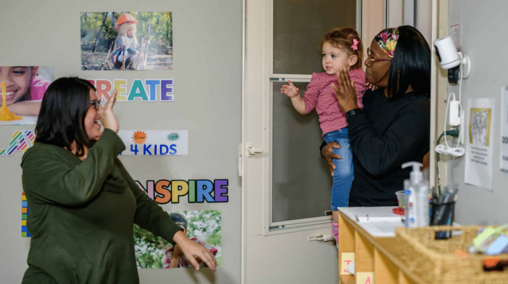 Woman holding a toddler greets another woman waving in a colorful classroom setting with posters on the wall.