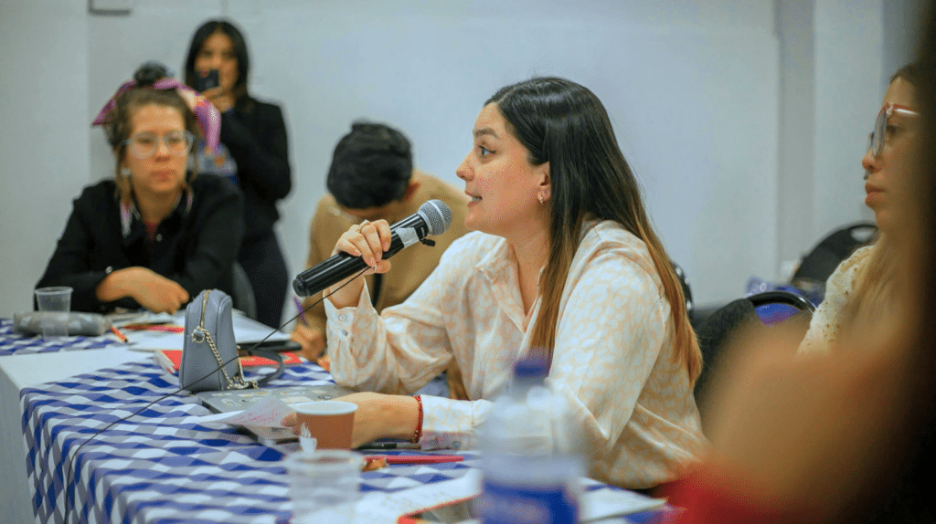 Woman in patterned shirt speaks into a microphone at a meeting, others listen around a checkered table.