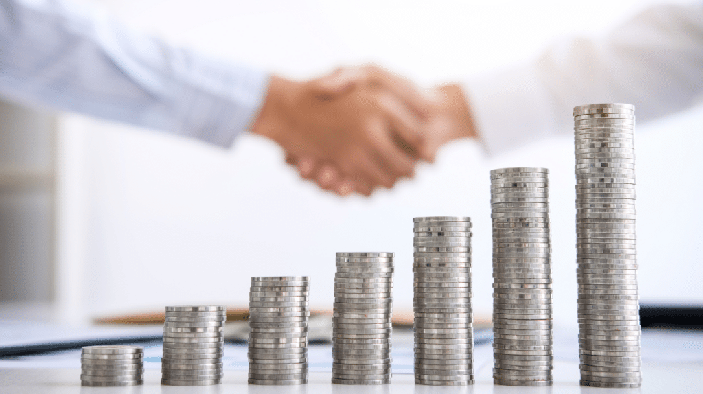 Stacks of coins in ascending order with two people shaking hands in the blurred background.