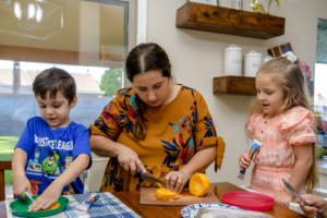 Woman chops bell pepper while two children prepare food at a kitchen table, all focused on their tasks.