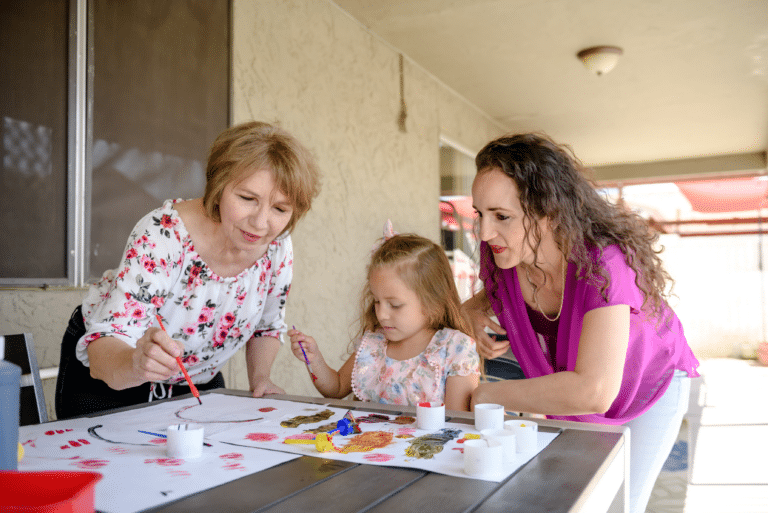 Three people paint together at a table outdoors, using brushes and colorful paints on white paper.