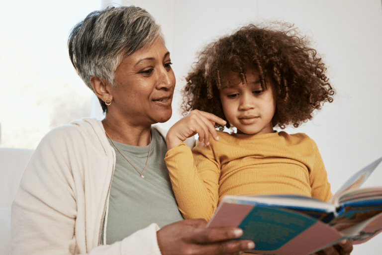 Older woman reading a colorful book to a young girl with curly hair, both sitting closely together.