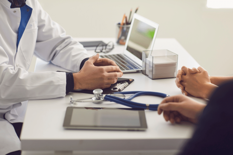 Doctor and patient sit across from each other at a desk with a laptop, stethoscope, and tablet.