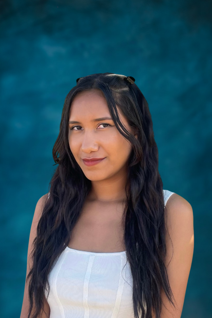Woman with long wavy hair in a white top, posing in front of a blue background.
