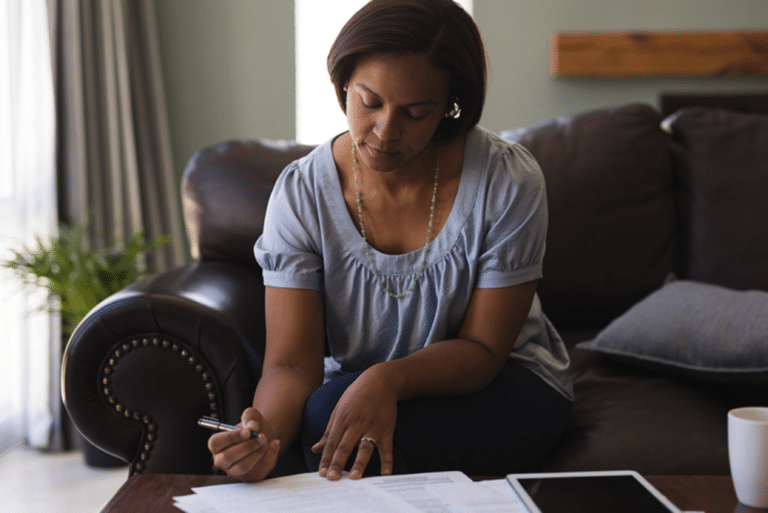Woman sitting on a couch, writing on papers with a pen, with a tablet, mug, and documents on the table.