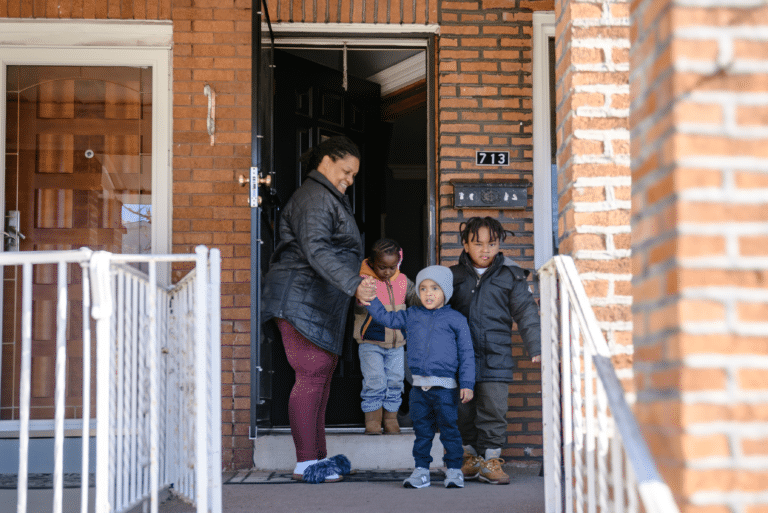 A woman and three children stand at the entrance of a brick house, preparing to leave together.