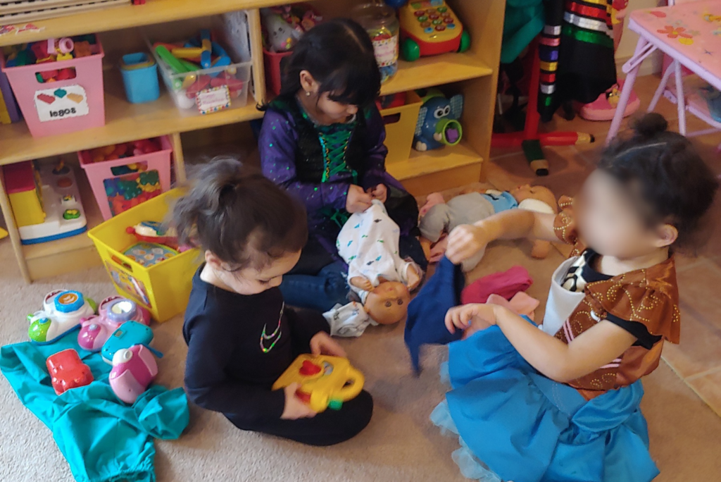 Three children sit on the floor playing with baby dolls and plastic toys.