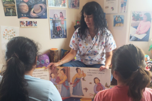 A woman in a patterned scrub top reads an open picture book to two children.