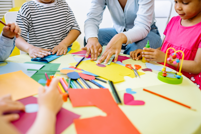 A teacher and three children work on a colorful paper craft project at a round table.