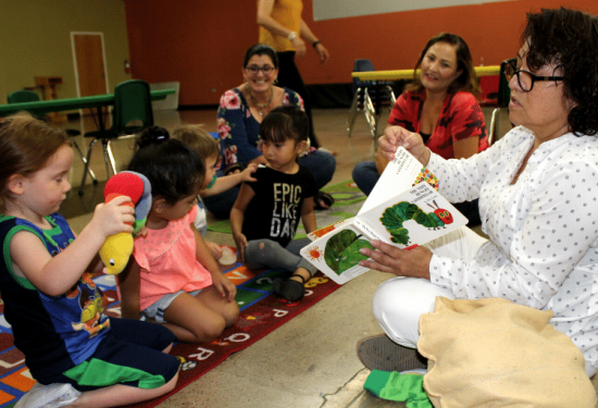 A woman reads a picture book to young children sitting on a colorful rug in a classroom.
