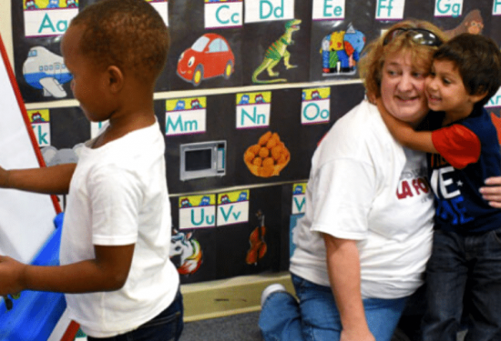 A young boy hugs a teacher while another child plays with magnetic letters in a classroom.