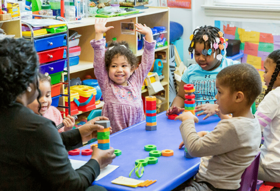 Children and a teacher play with colorful blocks at a classroom table, with one child excitedly raising her arms.