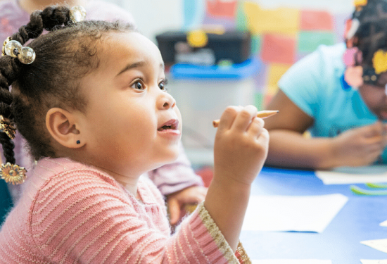 Young girl with braids and beads holding a pencil, sitting at a table, engaged in classroom activity.