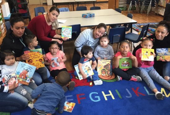 Adults and children sit on a colorful alphabet rug, holding picture books and smiling in a classroom setting.