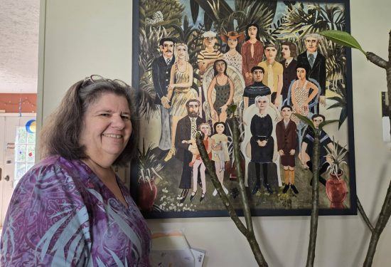 Smiling woman in a purple shirt stands by a colorful painting of many people in a garden setting.
