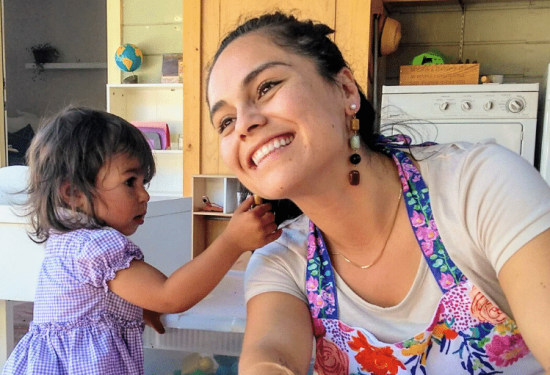Smiling woman in a colorful apron with a young girl indoors near a washing machine and wooden shelves.