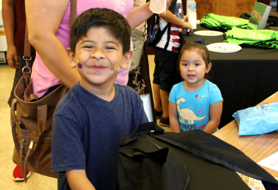 Smiling boy with stickers on his face stands by a table, with a girl in blue shirt behind him indoors.