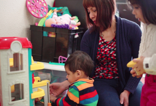 Woman and two children play with a toy house; toys and a doll are visible in the background.