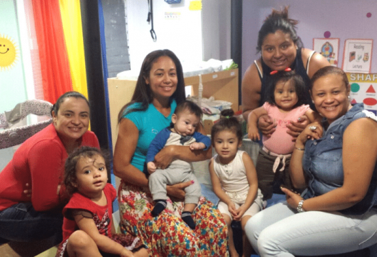 Five women and four young children smiling together in a colorful playroom with educational posters on the wall.
