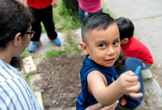 Young boy outdoors holds up a crayon, looking at the camera, with other children and an adult nearby.