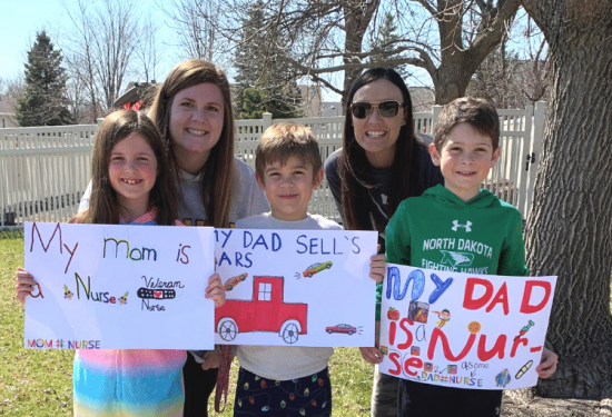 Five people smile outdoors, holding handmade signs about their parents' jobs as nurse and car salesperson.