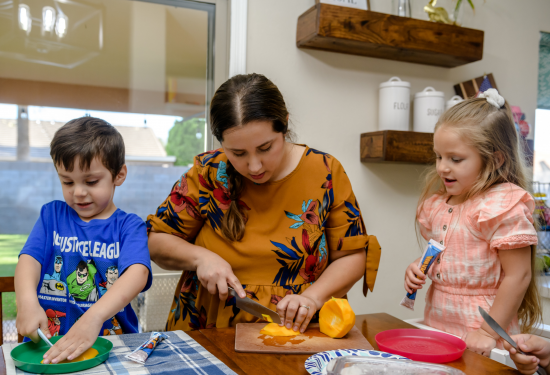 Woman chops bell pepper while two children prepare food at a kitchen table, all focused on their tasks.