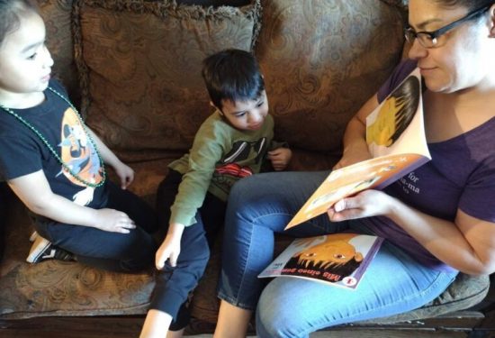 A woman reads a book to two children sitting with her on a patterned couch.