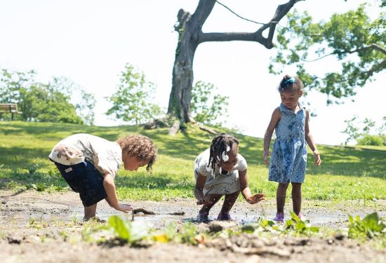Three young children play and explore in a muddy area in a sunny park with green grass and trees.