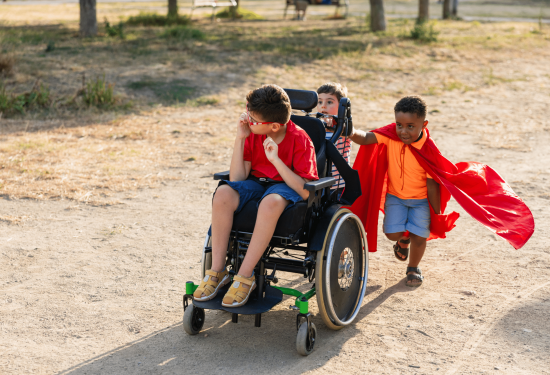 Three kids play outdoors, one in a wheelchair, while another pushes and both wear red capes.