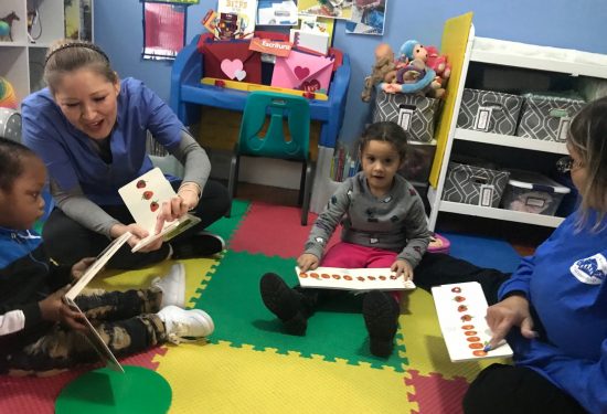 Two adults and two children read picture books together on a colorful play mat in a classroom setting.
