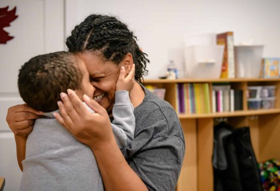 Adult and child smiling and hugging closely in a warm, joyful moment in a cozy room with bookshelves behind them.