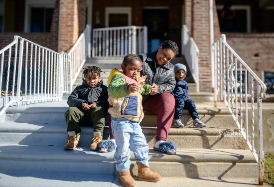 A woman and three children sit together on outdoor steps in front of a brick house on a sunny day.