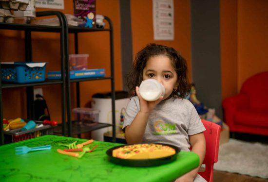 A young child sits at a green table, drinking from a bottle, with a plate of food in front of them.