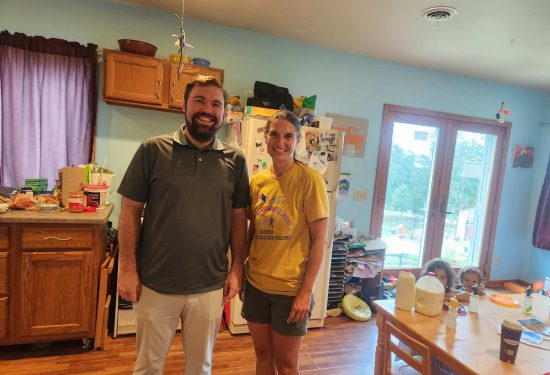 Two adults smile in a kitchen while children sit at a table in the background near a glass door.