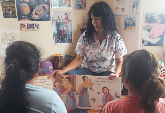 A woman in a patterned scrub top reads an open picture book to two children.