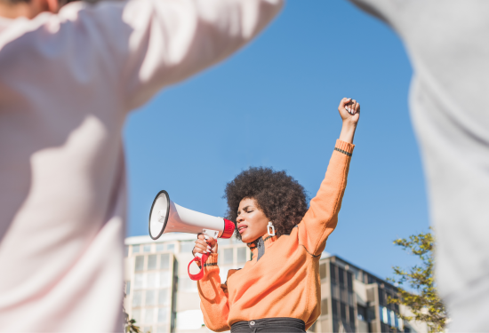 A woman with a megaphone raises her fist while protesting outdoors under a clear blue sky.