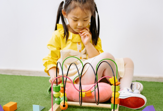 Young girl in yellow sits on grass, playing thoughtfully with a wooden bead maze and colorful blocks.