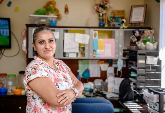 Woman in a floral shirt stands smiling with arms crossed in a colorful, cluttered home office space.