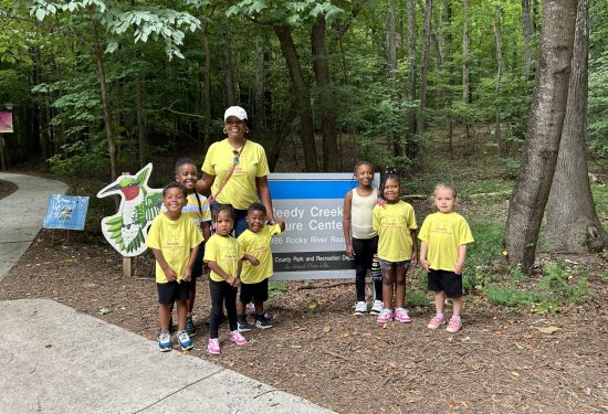 A group of kids and an adult pose by a "Reedy Creek Nature Center" sign in a wooded area.