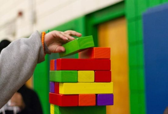 A hand reaches to place a green block on top of a colorful stacked block tower indoors.